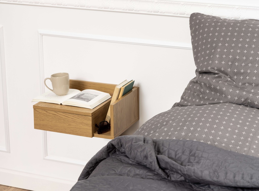 Oak bedside table with one drawer, featuring a minimalist design, complemented by a coffee cup, books, and sunglasses on top, set against a textured gray bedding backdrop.