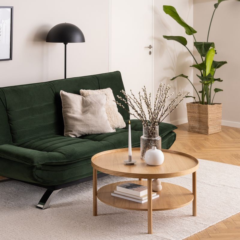 Round oak coffee table with a natural finish, featuring a lower shelf, placed in a cozy living room setting with a green sofa, decorative items, and a potted plant.