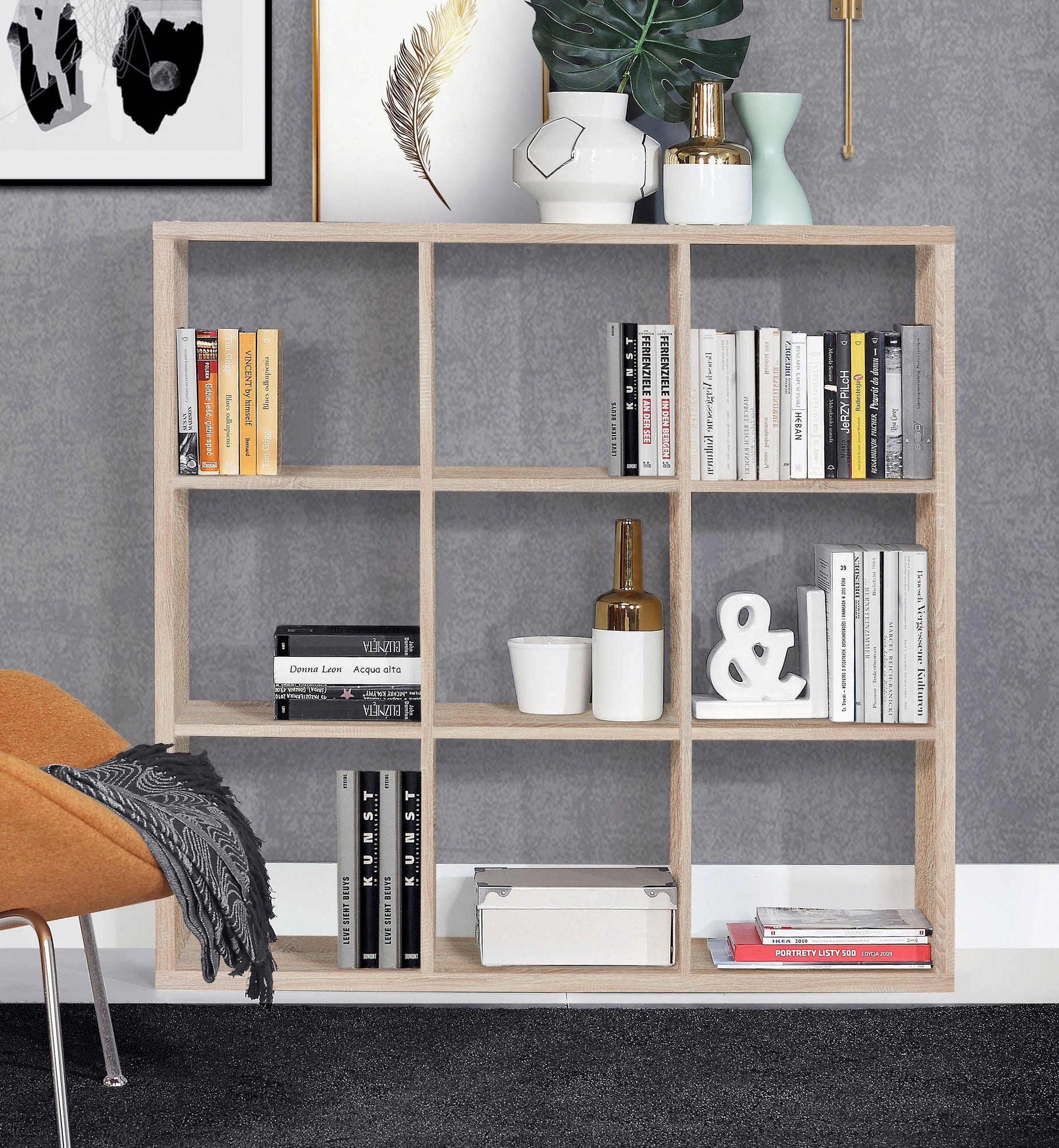 Mauro 3X3 Storage Unit in Sonoma Oak features a light brown finish, open shelving design, and is styled with books, decorative vases, and a modern chair against a textured gray wall.