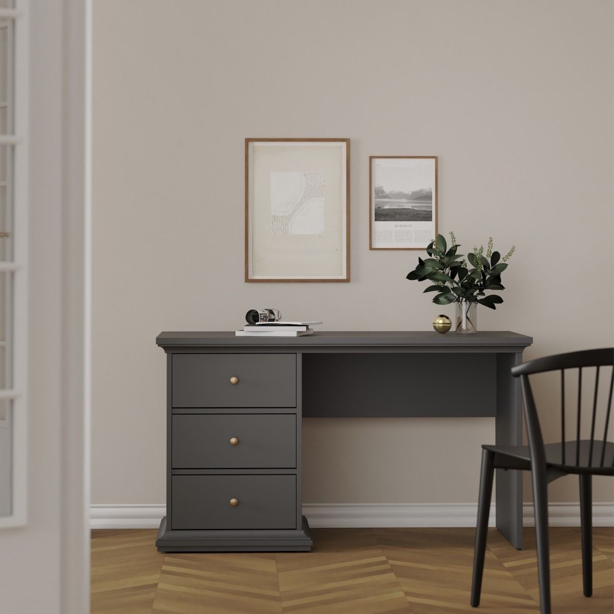 Matt grey wooden desk with three drawers, gold knobs, and a minimalist design, complemented by a black chair and decorative wall art in a neutral room setting.