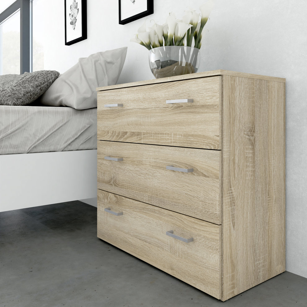 Oak effect chest of three drawers in brown, featuring sleek silver handles, positioned beside a bed with a decorative vase of flowers on top.