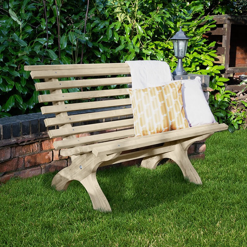 Curved wooden garden bench in natural finish, featuring slatted backrest and seat, with two decorative cushions, set against a lush green garden backdrop.