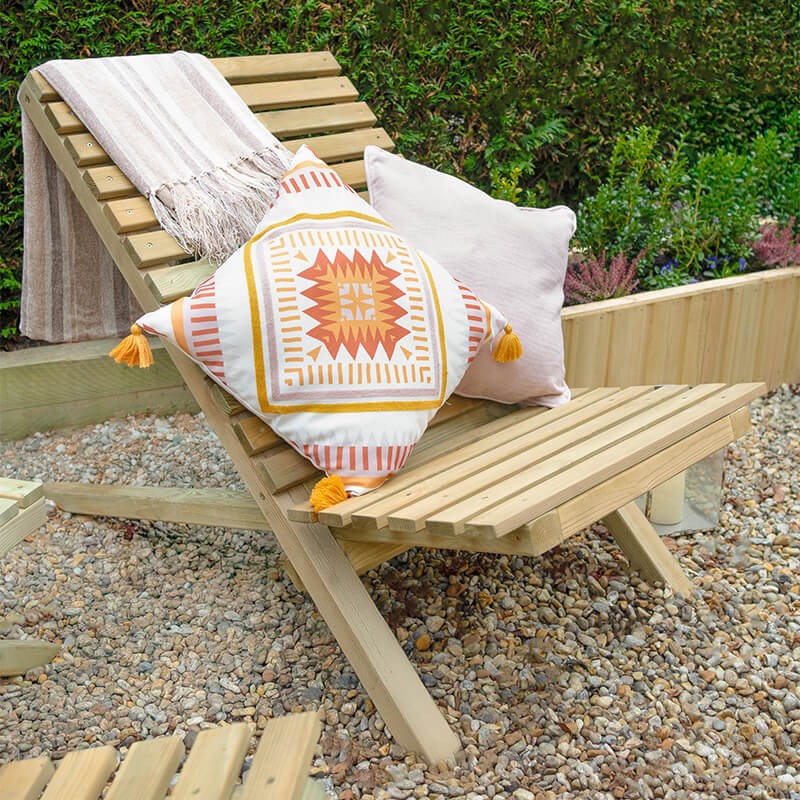 Slatted wooden garden chair in natural wood finish, featuring a reclining backrest, accompanied by a patterned cushion and a soft throw draped over it, set in a gravel garden area.