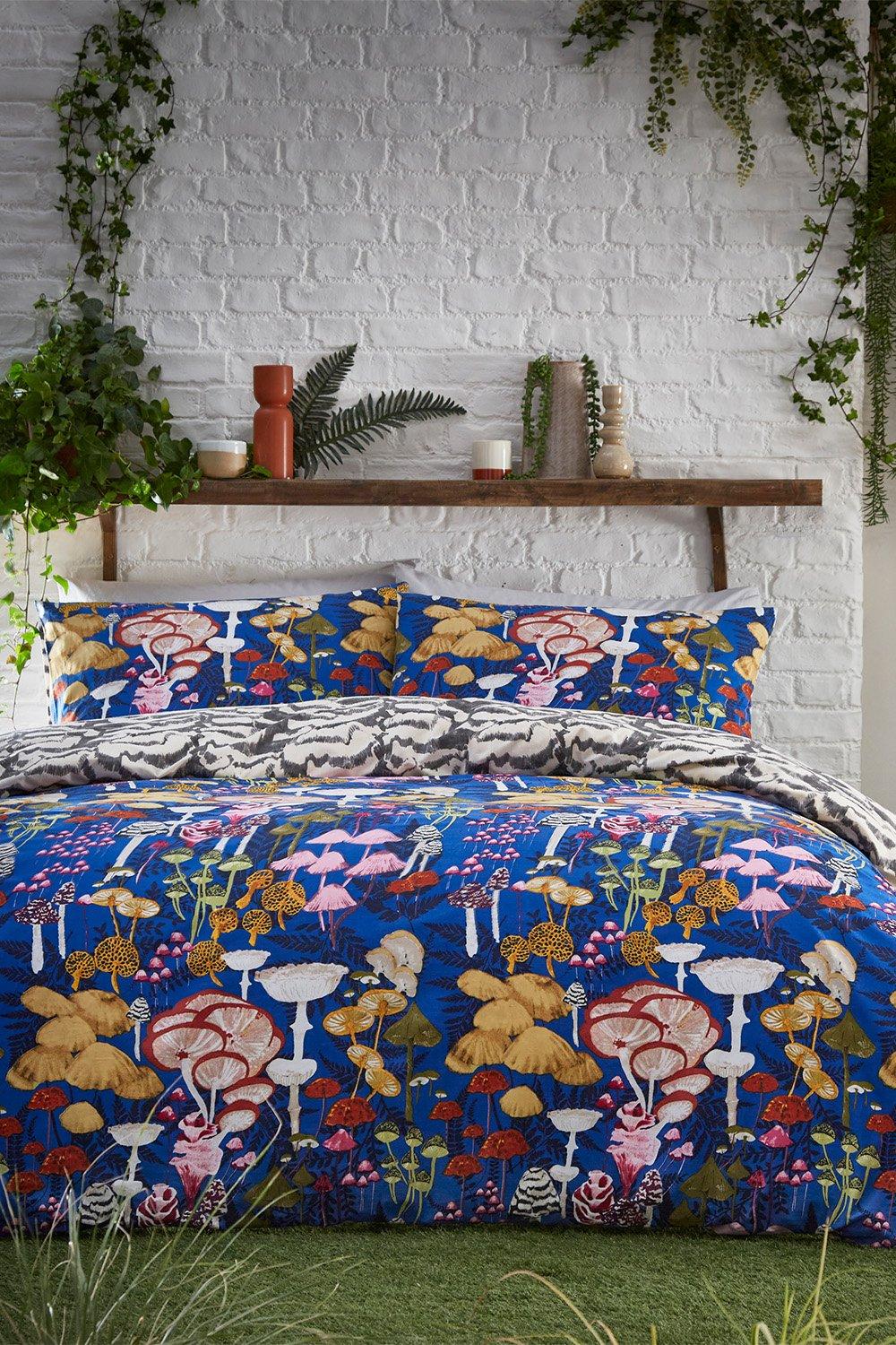 Cobalt blue duvet cover set featuring vibrant abstract mushroom designs in various colors, complemented by matching pillow shams, set against a textured white brick wall.