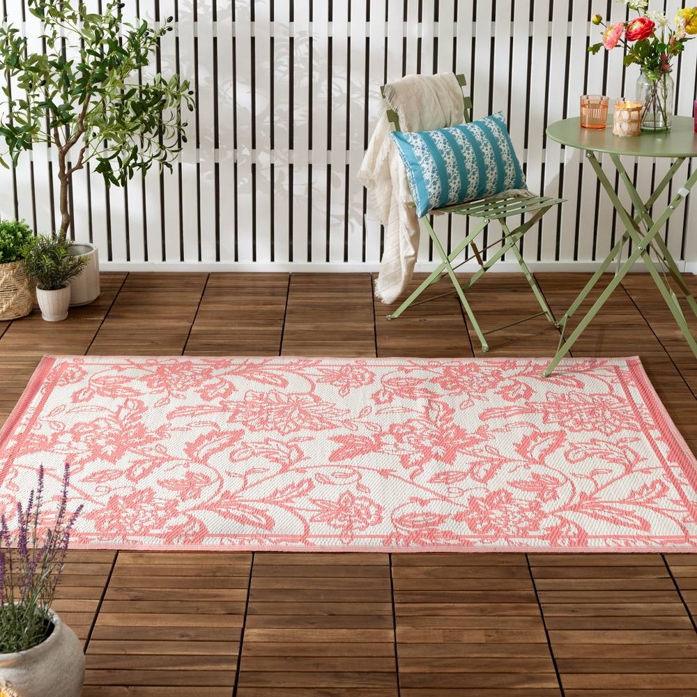 Woven jacquard outdoor rug featuring a floral pattern in coral and white, set on a wooden deck with green furniture and potted plants.