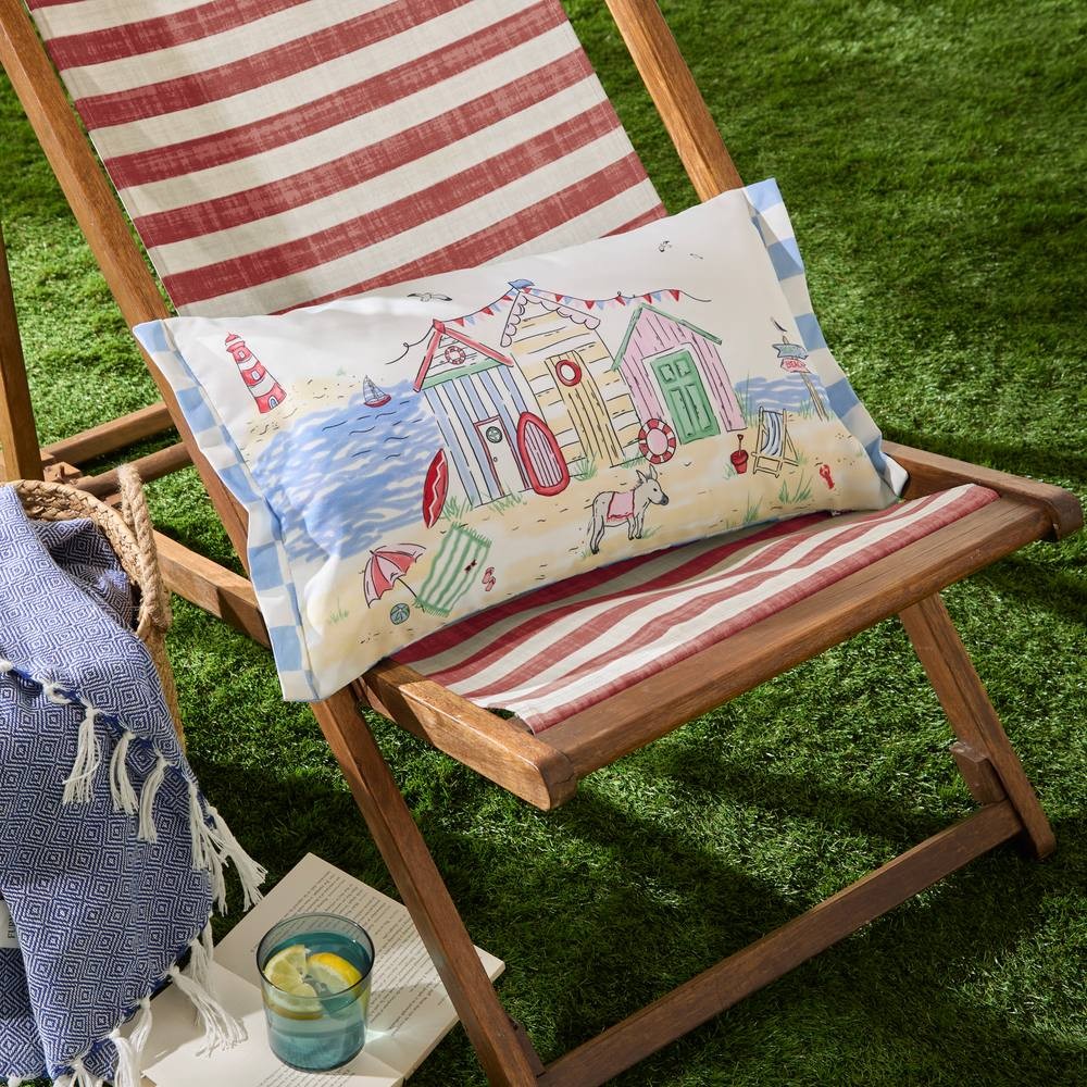 Rectangular outdoor cushion featuring colorful beach huts, made from durable fabric, with a playful seaside design, placed on a striped deck chair beside a drink and a blanket.