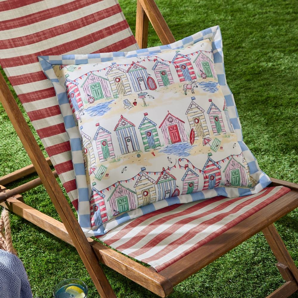 Square outdoor cushion featuring a colorful beach hut design with red, green, and blue houses, bordered by a light blue check pattern, set against a grassy background.