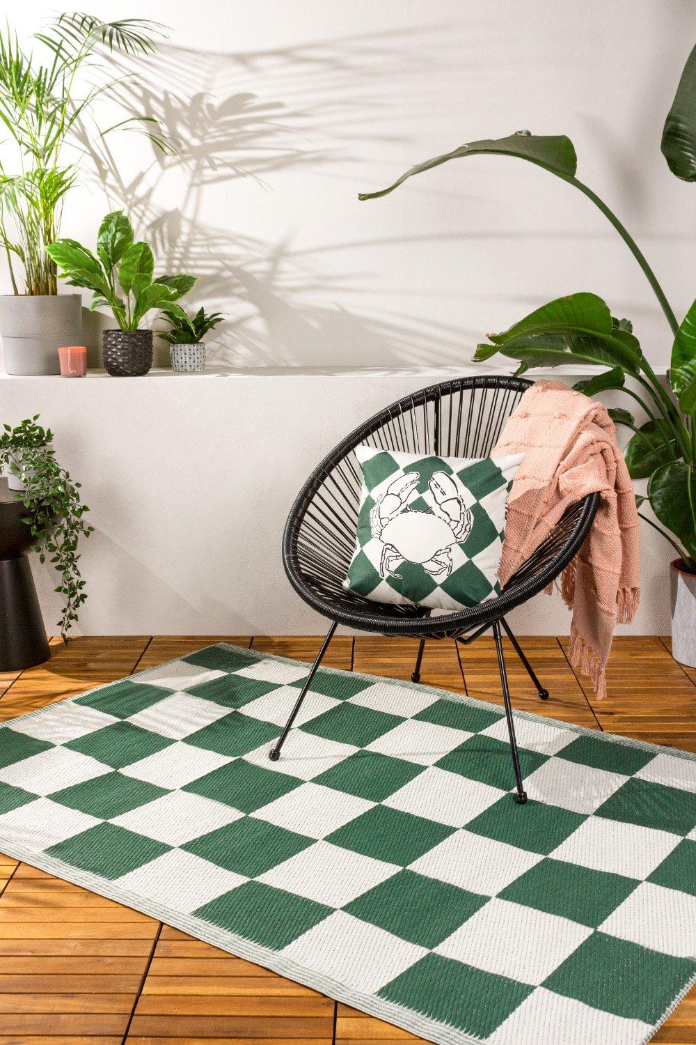 Green and white checkerboard outdoor rug laid on wooden flooring, accompanied by a black wire chair, decorative pillow, and a soft pink throw, with lush plants in the background.