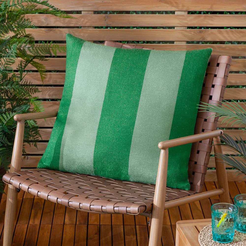 Green woven outdoor cushion featuring bold, alternating light and dark stripes, displayed on a wooden chair amidst greenery and decorative glasses.