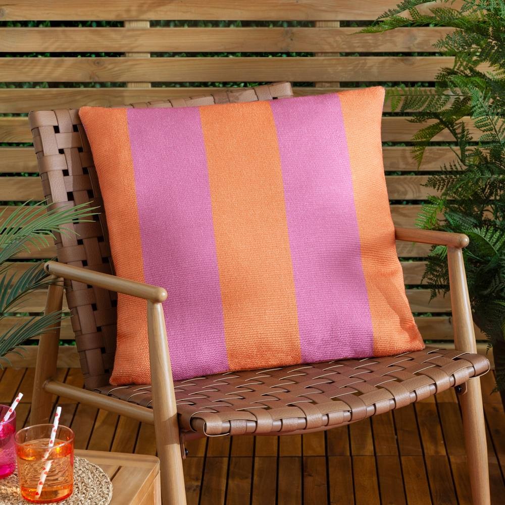 Orange and pink striped woven outdoor cushion, featuring a textured fabric, placed on a wooden chair surrounded by greenery and colorful drinks.