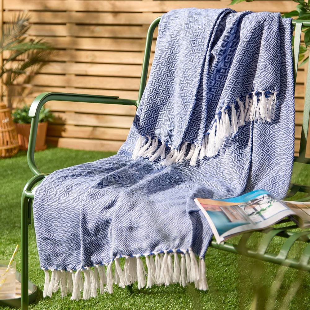 Cobalt blue woven throw with a geometric pattern and white fringes, draped over a green metal chair in a sunny outdoor setting.