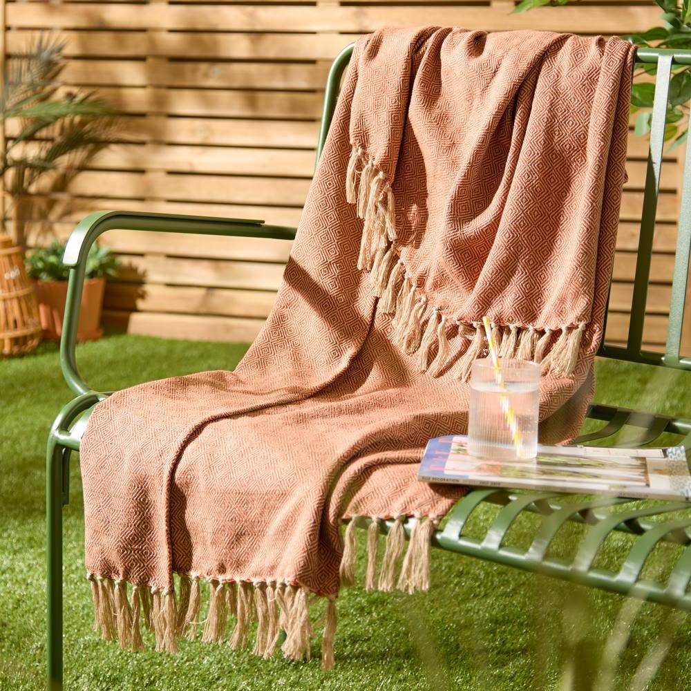 Terracotta woven throw with geometric patterns and fringed edges, draped over a green metal bench, accompanied by a glass of drink and magazines on a grassy outdoor setting.