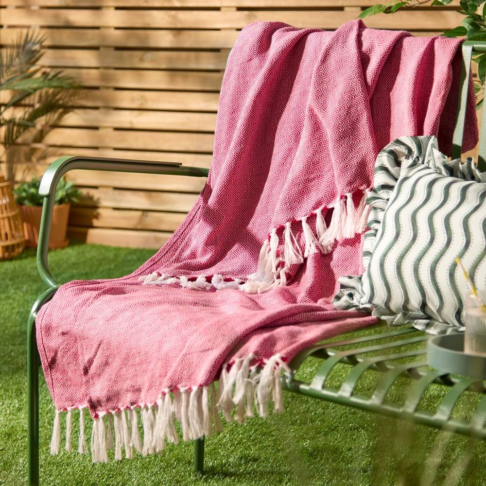 Woven pink throw blanket with white fringes draped over a green metal chair, complemented by a patterned cushion, set against a wooden backdrop and lush greenery.