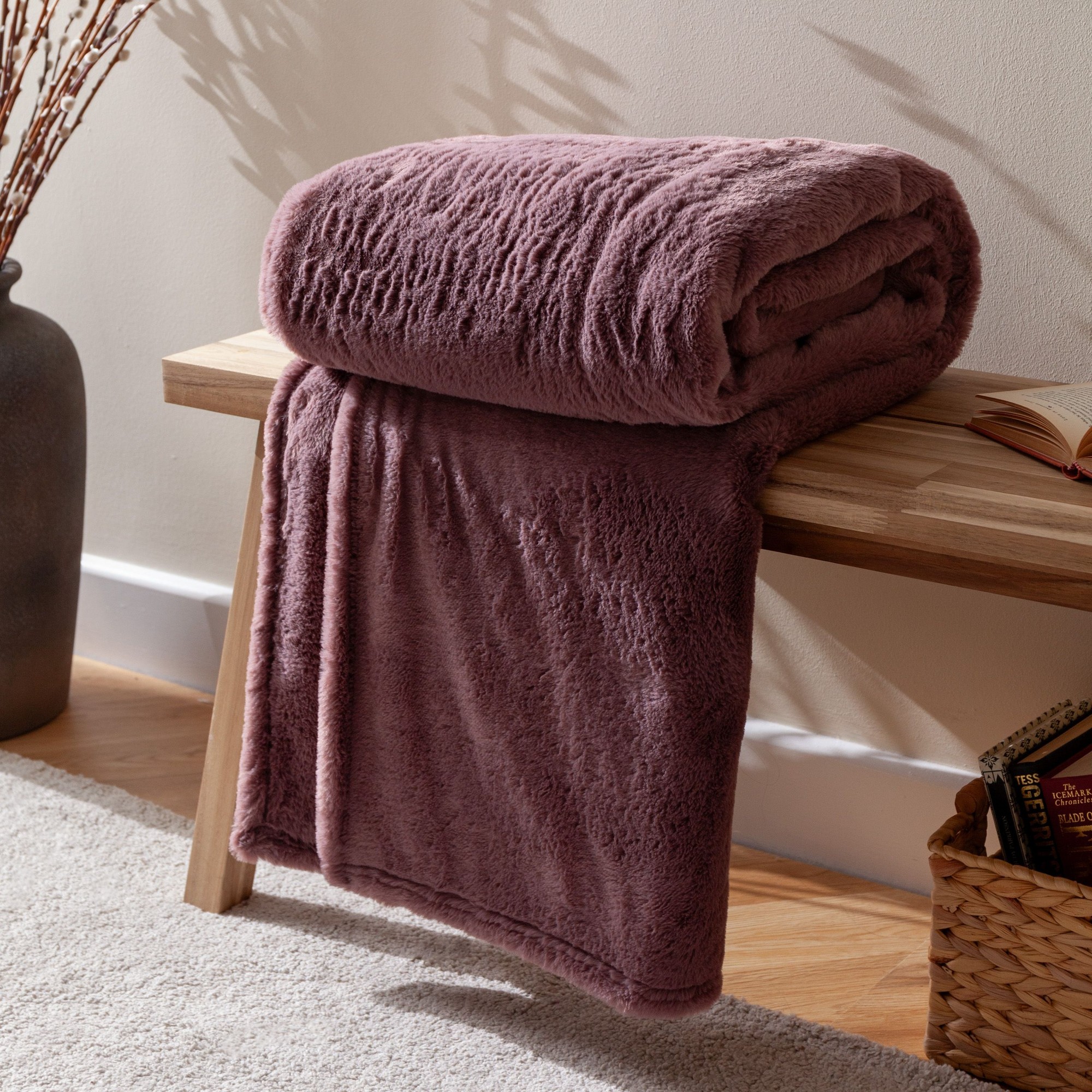 Soft double-sided faux-fur throw in a muted mauve color, measuring 150cm x 200cm, neatly rolled and draped over a wooden bench with a textured surface.