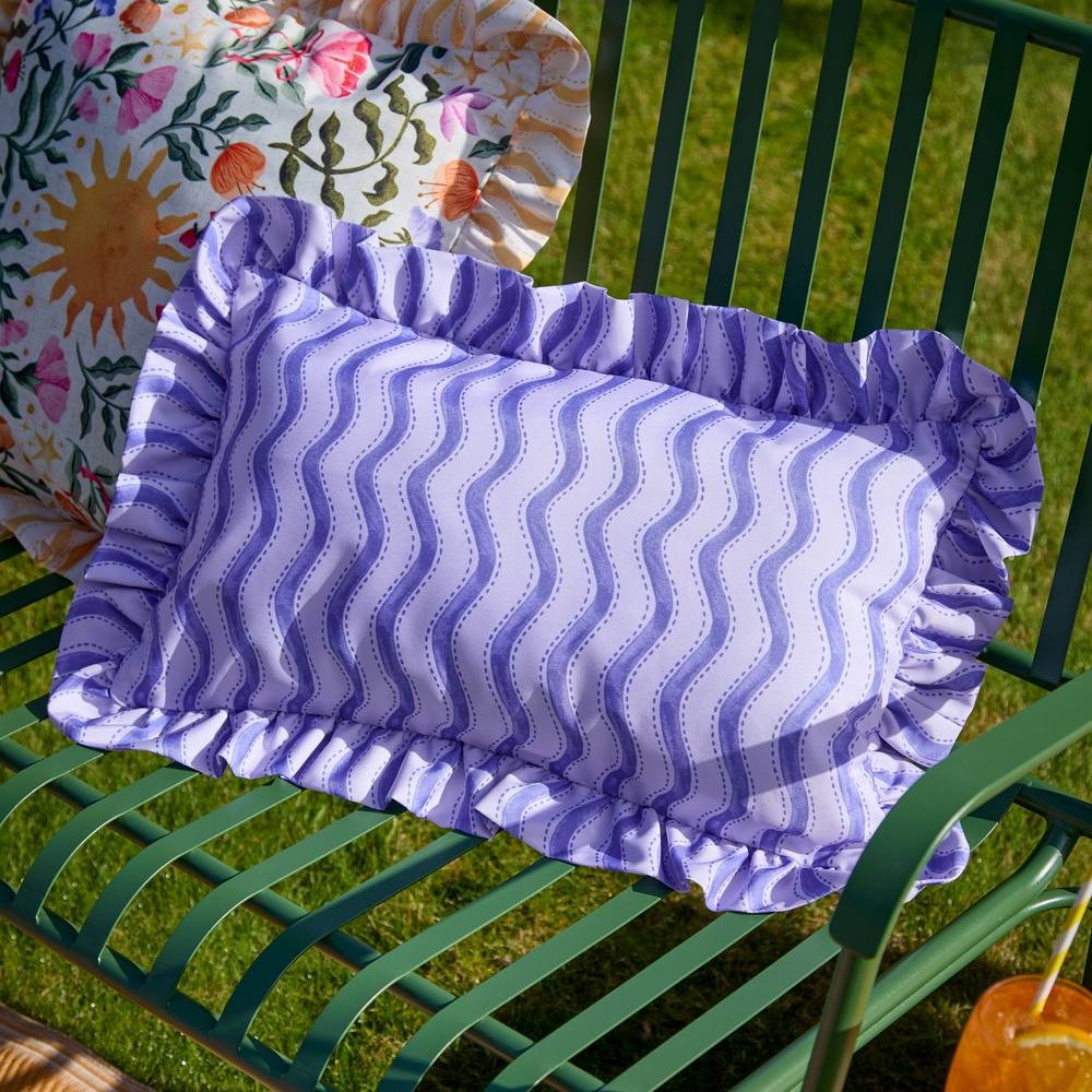 Violet outdoor cushion with wiggly stripe pattern and ruffled edges, placed on a green metal bench, surrounded by a floral cushion and grass.