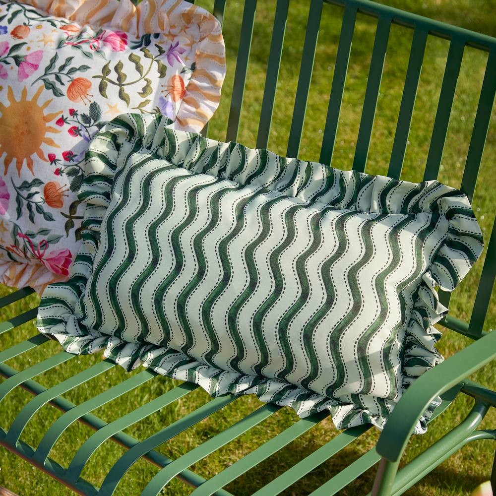 Green outdoor cushion with wiggly stripe pattern and ruffled edges, featuring a soft fabric, placed on a green metal bench in a grassy setting.