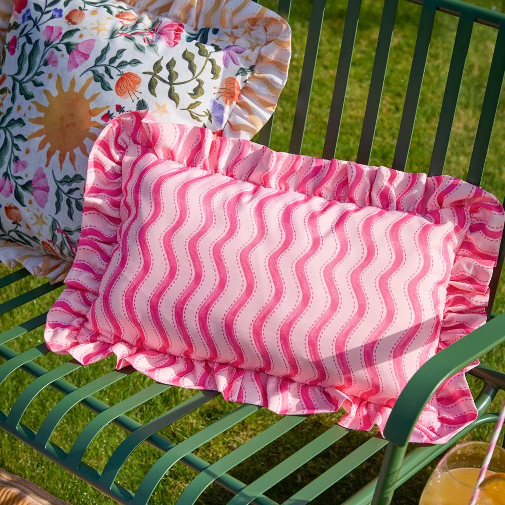 Rectangular outdoor cushion in pink with wavy stripe pattern, featuring ruffled edges, placed on a green metal chair, alongside a floral patterned cushion.