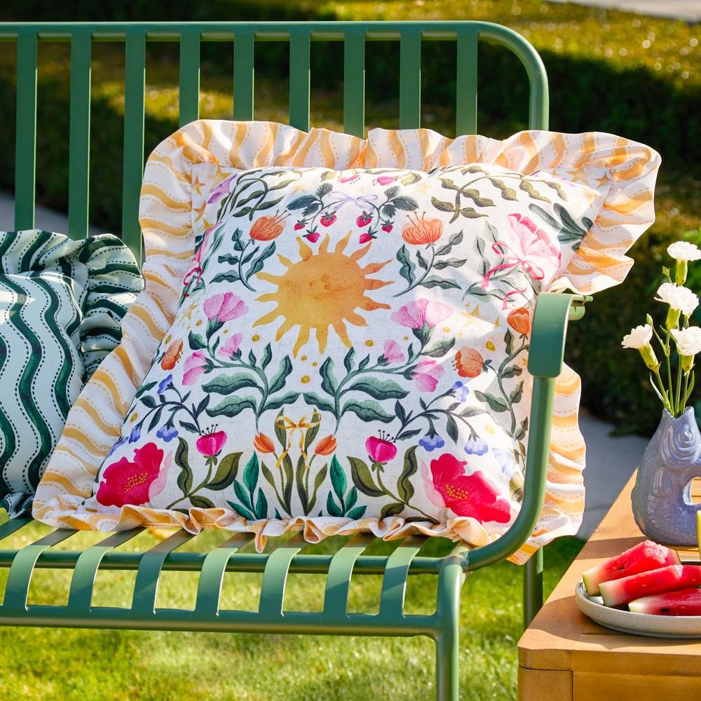 Colorful water-resistant outdoor cushion featuring a sun and floral design, with a ruffled yellow and white striped border, placed on a green metal bench.