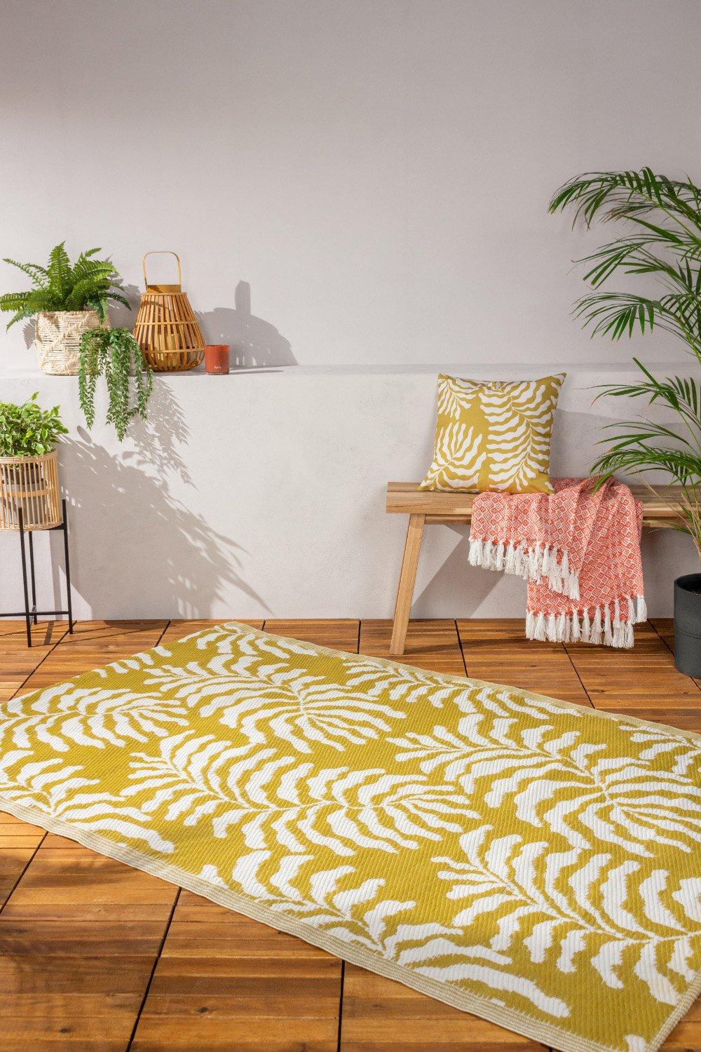 Mustard yellow woven rug featuring a white fern leaf pattern, set in a stylish indoor/outdoor space with plants and decorative accents.