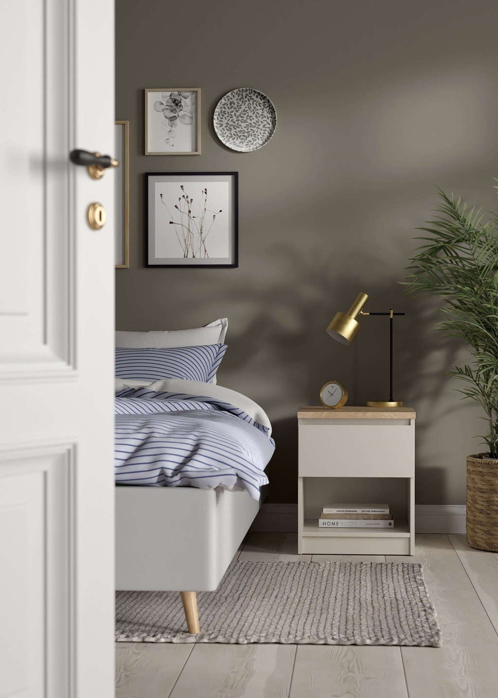 Naia bedside table in cashmere Jackson hickory features one drawer and one shelf, complemented by a gold lamp, decorative plates, and a potted plant in a cozy bedroom setting.