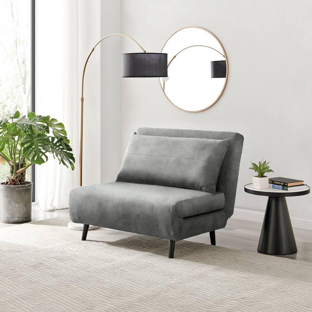 Compact grey futon sofa bed upholstered in soft velvet, featuring a sleek design, two back cushions, and black wooden legs, positioned beside a modern floor lamp and a round mirror.