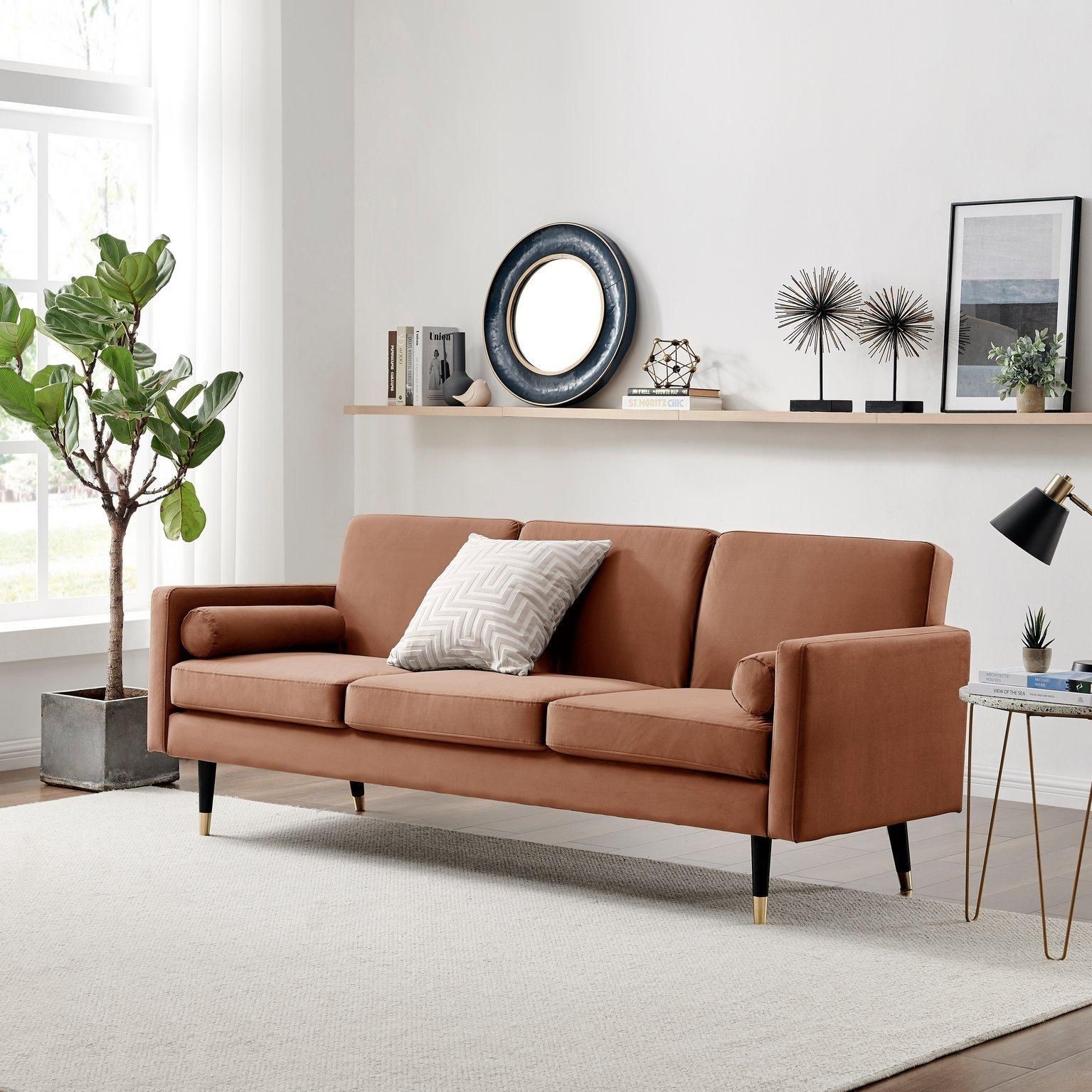 Brown velvet 3-seater futon-style sofa bed with sleek arms, tapered black legs, and a decorative cushion, set against a light wall and simple decor.
