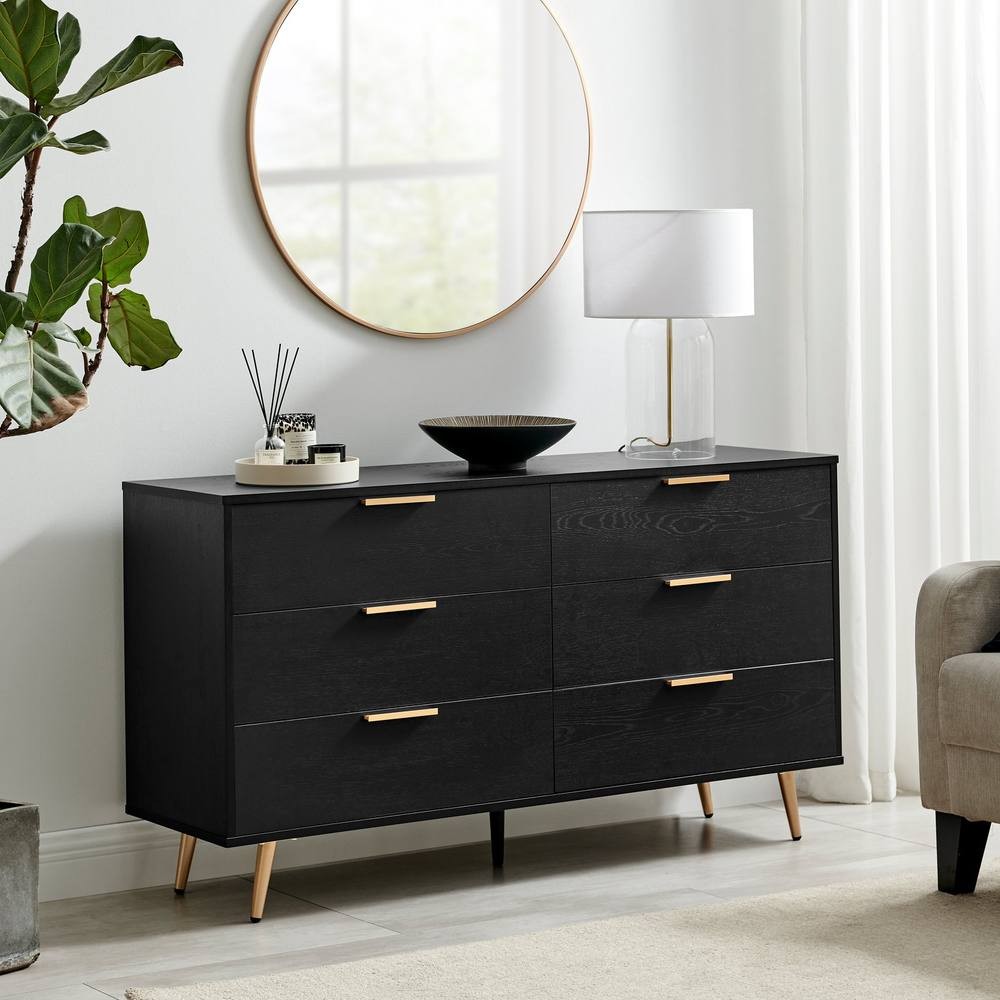 Black wooden chest of six drawers with copper effect handles, featuring tapered legs, a round mirror above, a lamp, and decorative items on top, set in a modern living space.