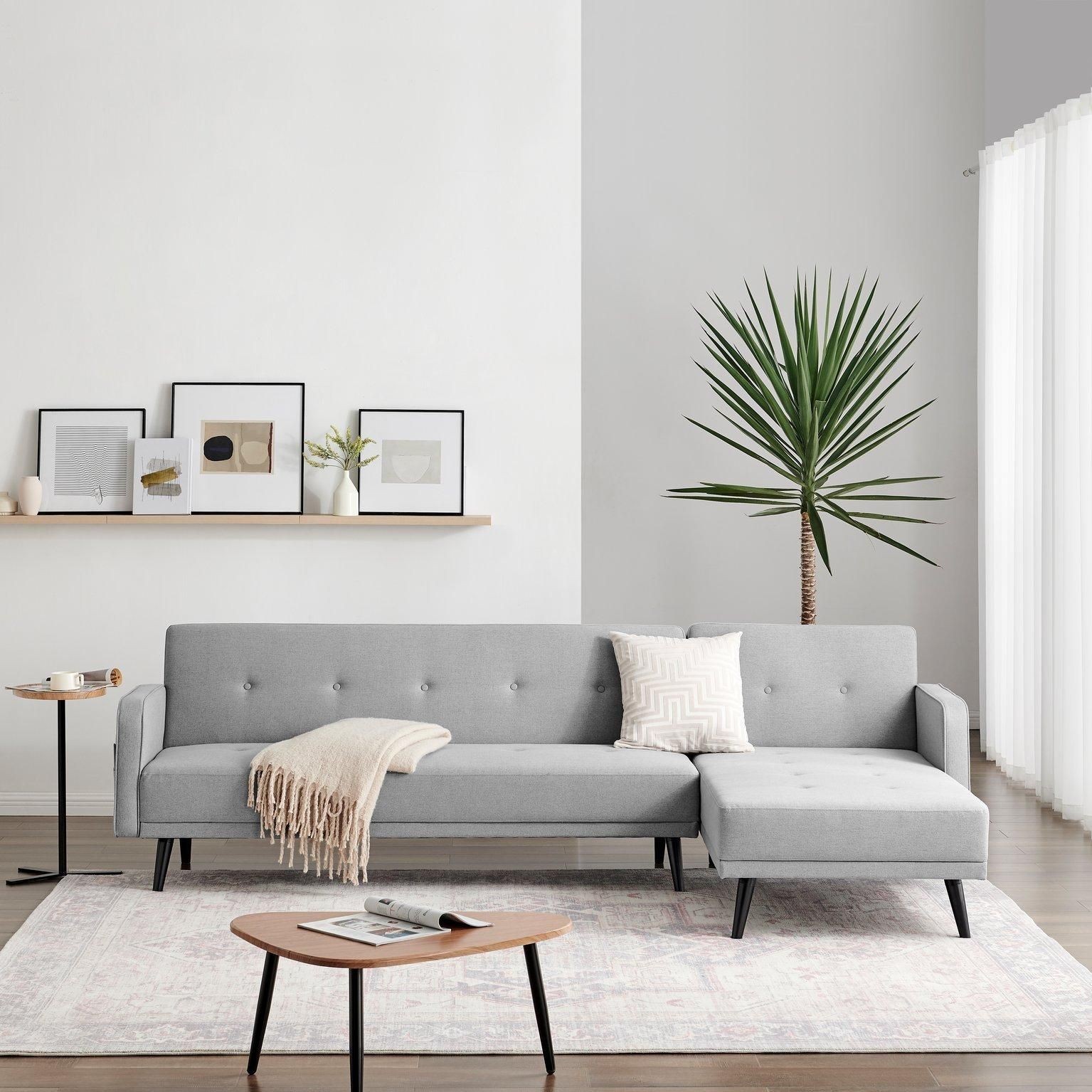 Grey fabric 3-seater L-shaped sofa bed with a click-clack mechanism, featuring tufted detailing, wooden legs, and a cozy throw pillow, set in a modern living space.