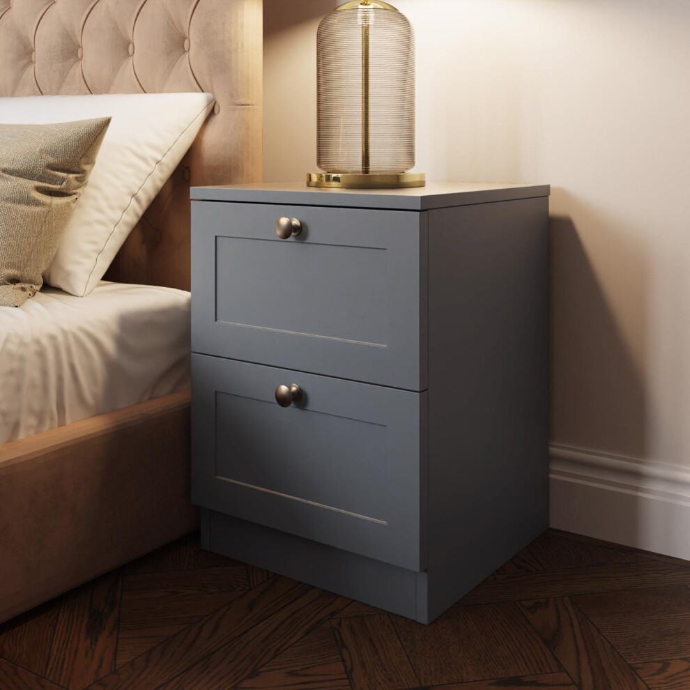 Dark grey panelled bedside chest table with two drawers, featuring round brass knobs, placed next to a bed with a tufted headboard and a decorative lamp on top.