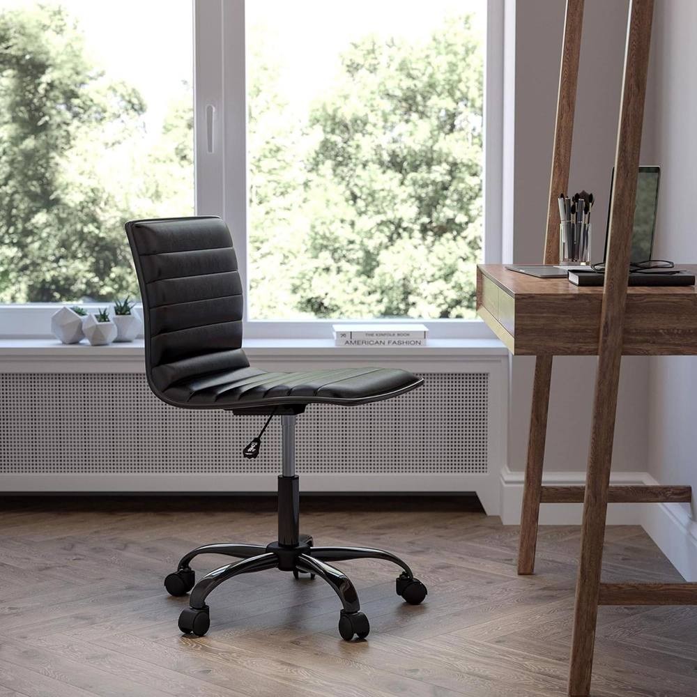 Black faux leather low back office chair with a ribbed design, adjustable height, and five rolling wheels, positioned beside a wooden desk in a bright room.