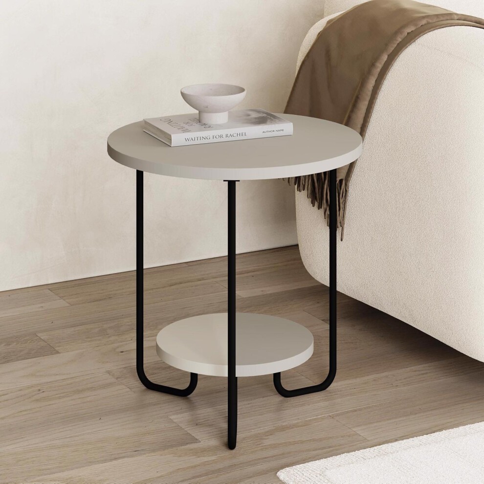 Beige, two-tier round side lamp table with a sandstone finish, featuring a sleek black metal frame and a lower shelf, styled with a small bowl and books on top.