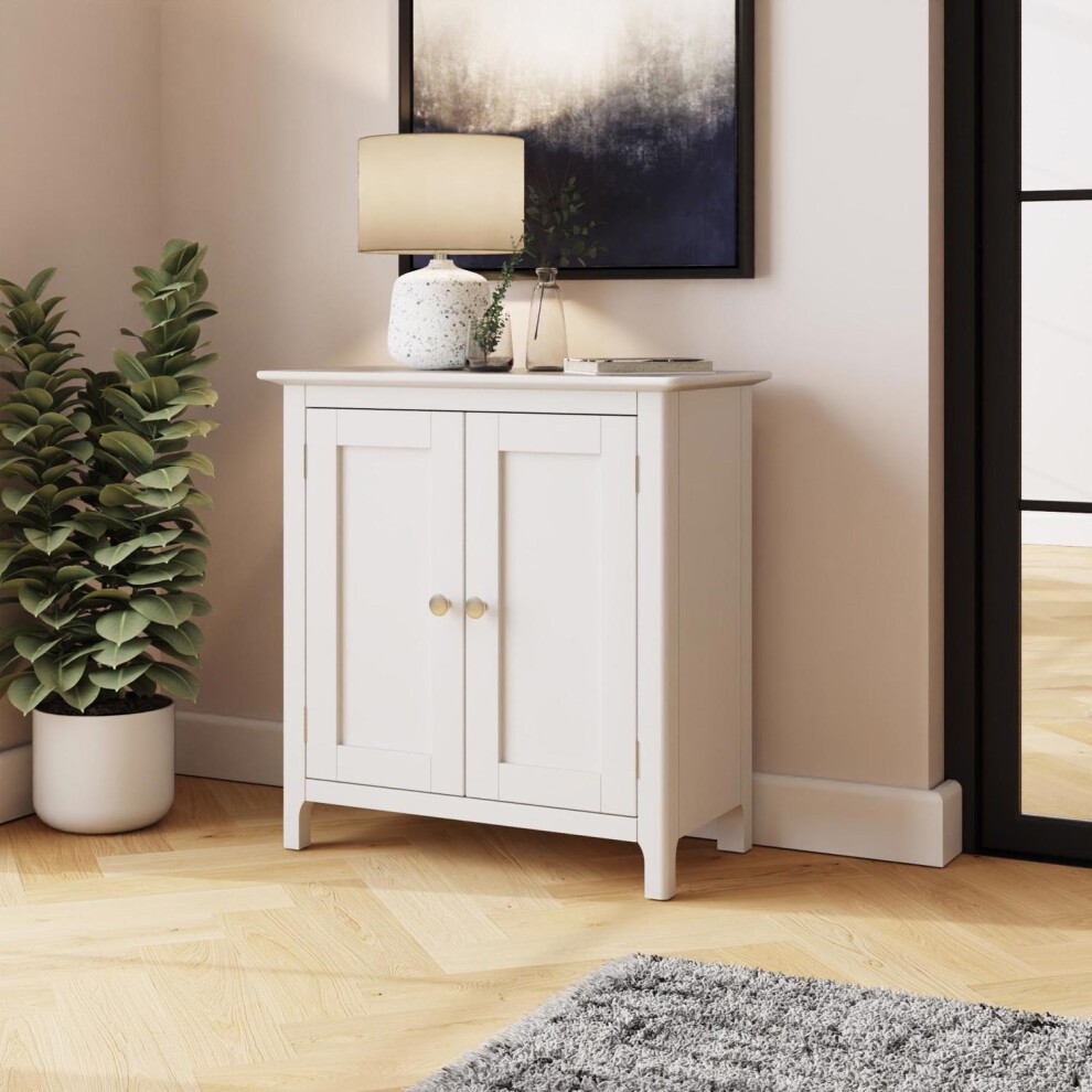 White compact sideboard with two doors, featuring round gold knobs, set against a light wall, accompanied by a lamp and a potted plant, on a wooden floor.