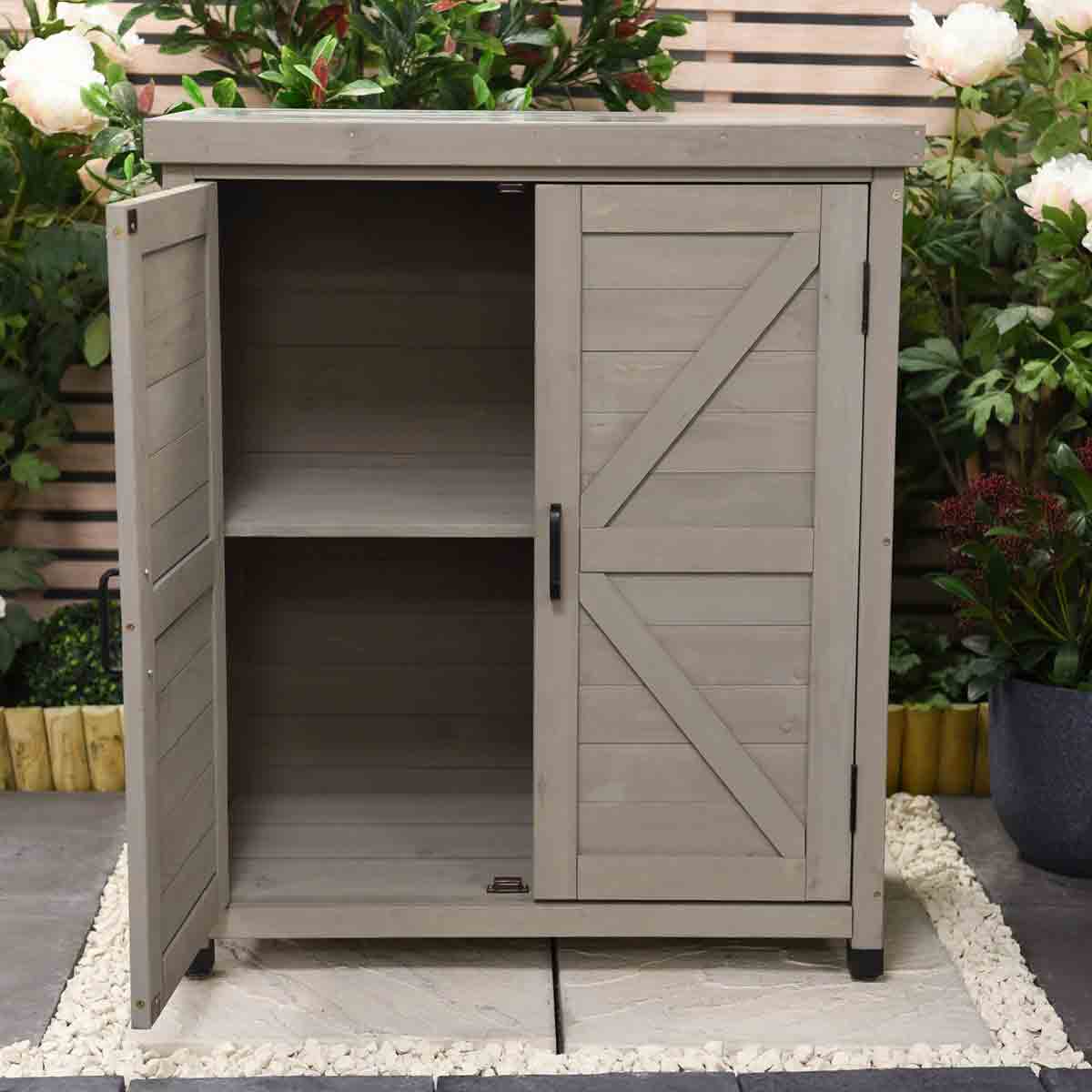 Grey wooden storage cabinet with a slatted design, featuring double doors and a black handle, set against a garden backdrop with plants and decorative stones.