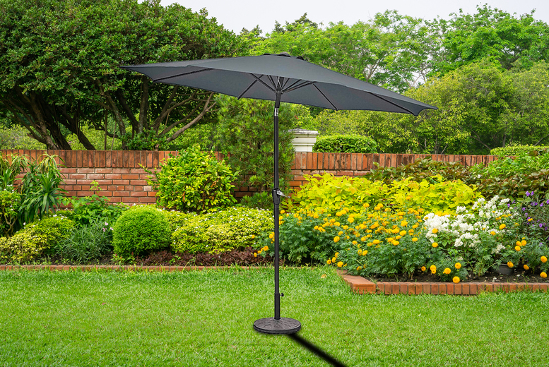 A charcoal-colored 2M parasol with a crank and tilt mechanism, featuring six ribs, stands in a lush garden with vibrant flowers and greenery.