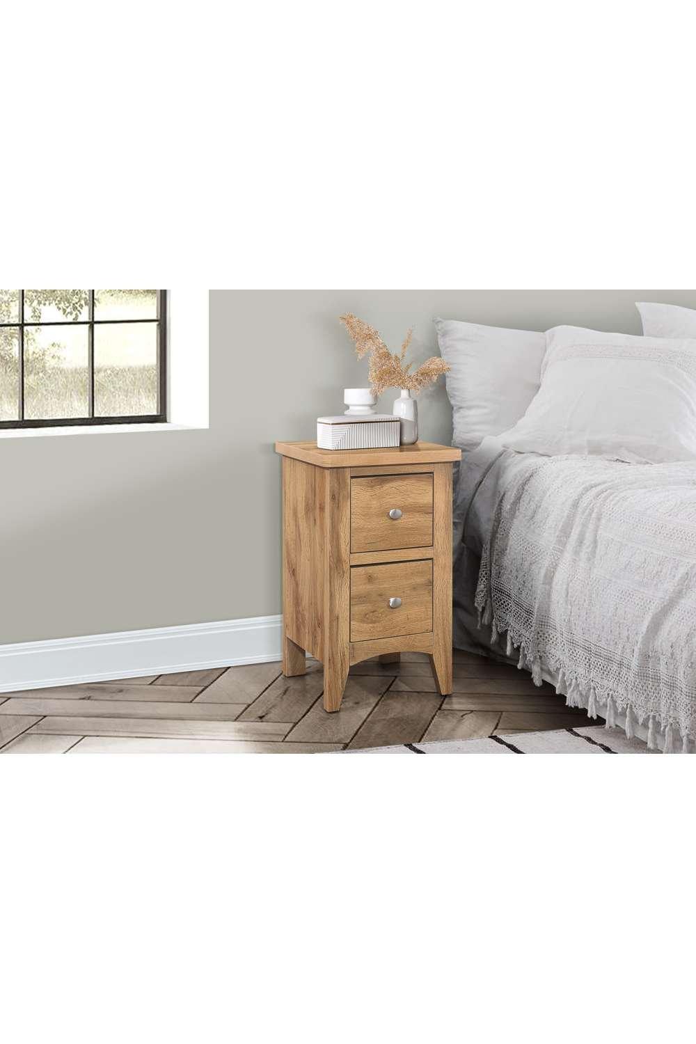 Brown oak bedside table with two drawers, featuring silver knobs, positioned next to a white bed with a textured blanket and a decorative vase on top.
