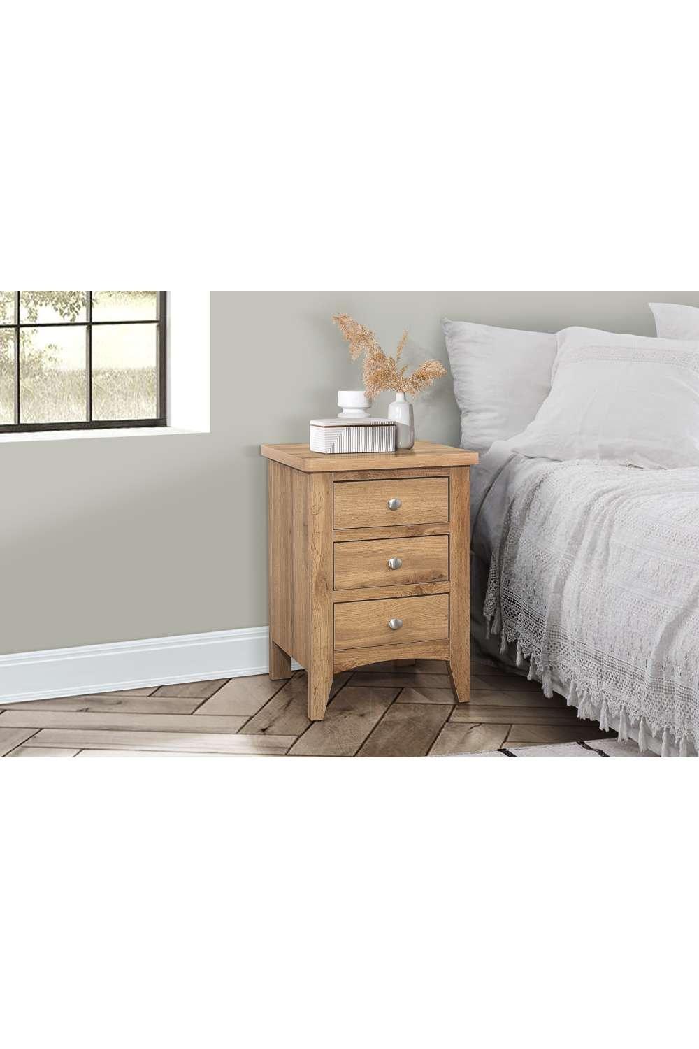 Brown wooden bedside table with three drawers, featuring silver knobs, set beside a bed with white bedding and decorative plants on top, against a light gray wall.