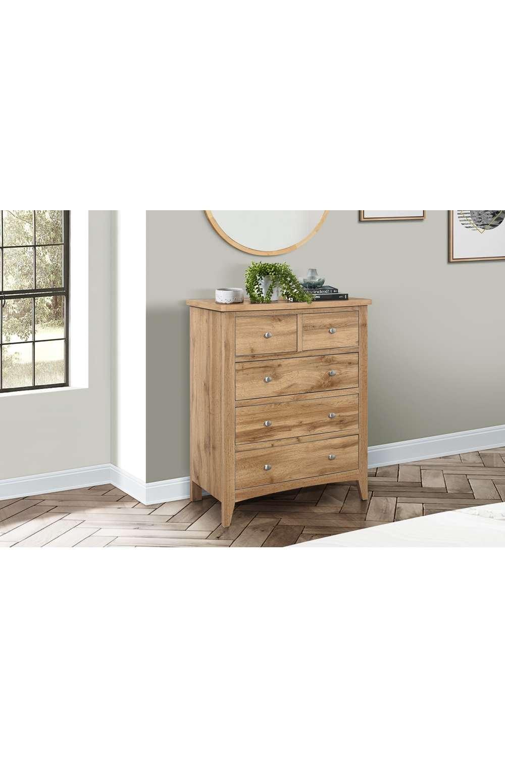 Birlea Hampstead 3 + 2 Chest in brown oak features a sleek design with five drawers and silver knobs, set against a light wall and wooden floor, complemented by decorative items.