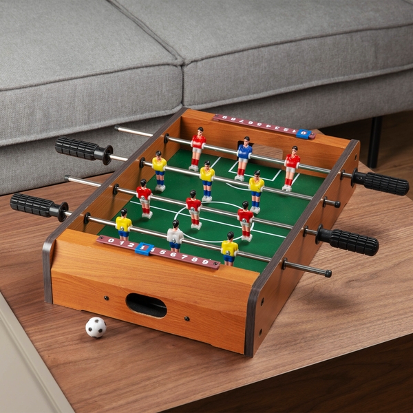 Table football set featuring a wooden base with a green playing field, plastic player figures in red and yellow, and black handles for control, placed on a modern couch.