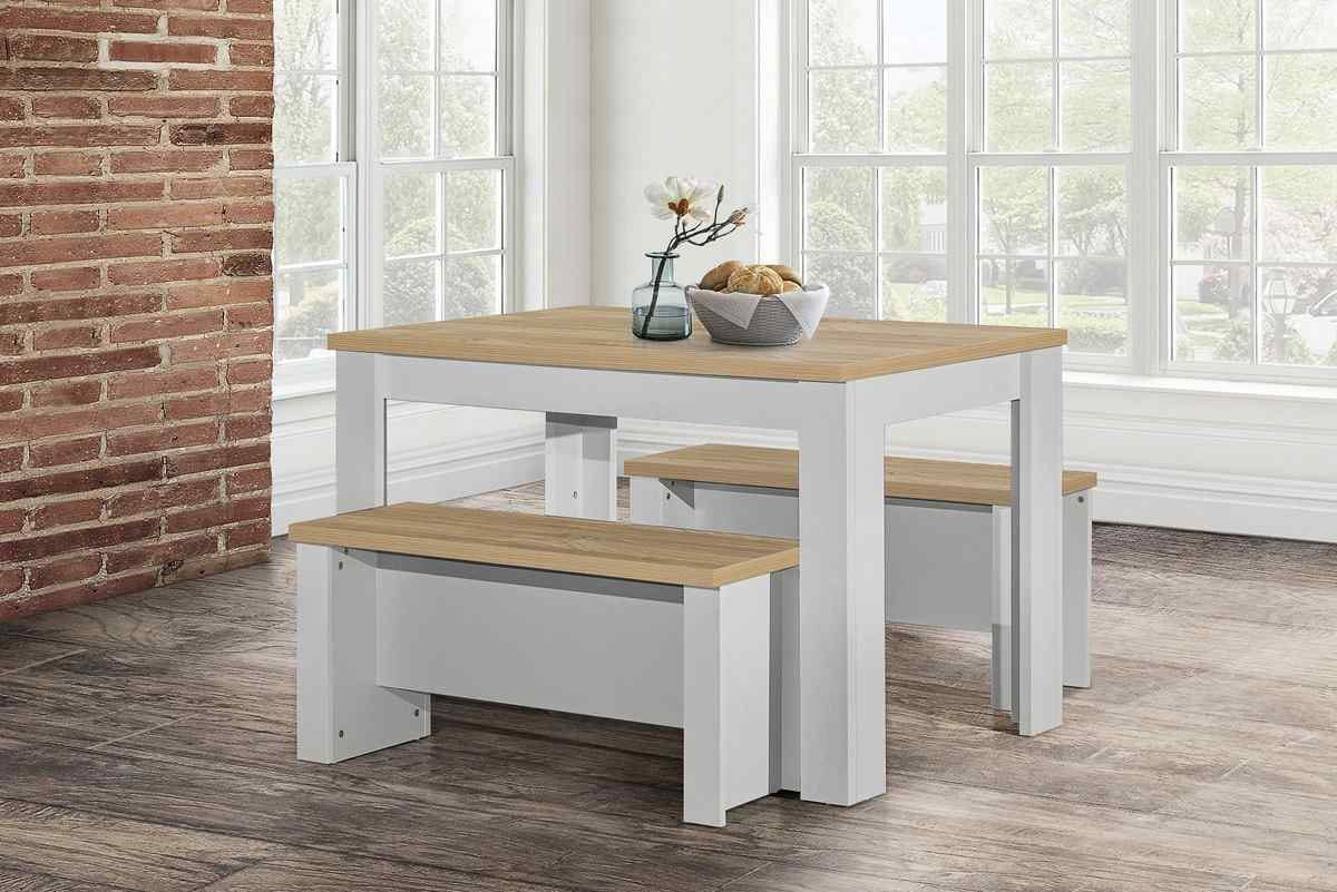 Grey traditional dining table and bench set with a light wood top, featuring a sturdy white frame, placed in a bright room with large windows and a brick wall.