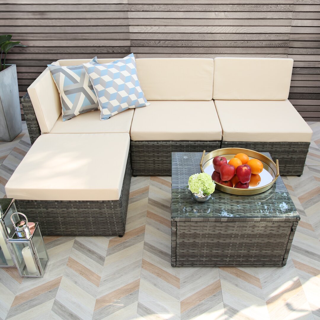 Lounge set featuring a modern design with a dark wicker finish, cream cushions, geometric patterned pillows, and a glass-top table adorned with fruit and a decorative plant.