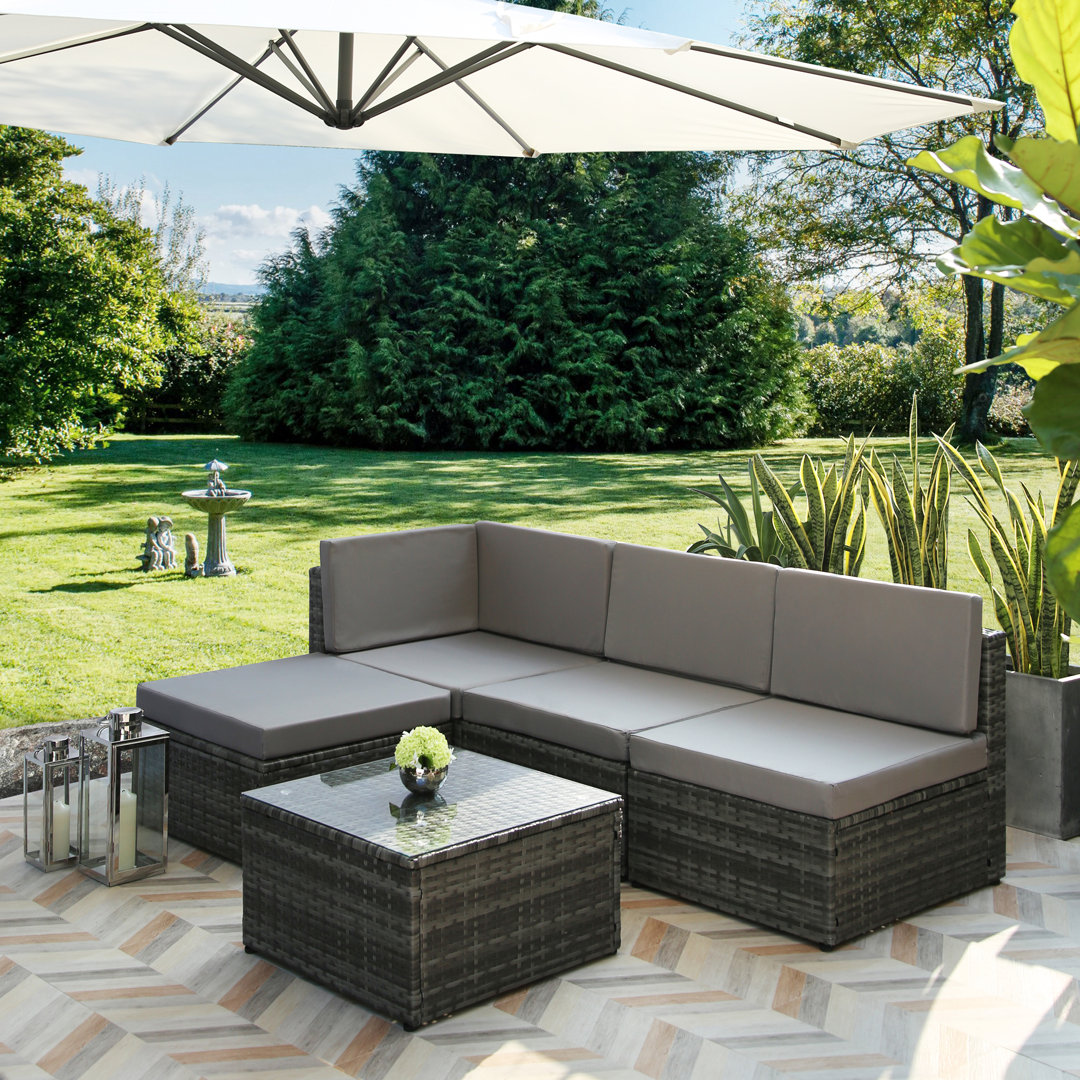 Stylish 3-seater lounge set in dark wicker with plush grey cushions, accompanied by a glass-top coffee table, set on a patterned patio under a large umbrella in a lush garden.