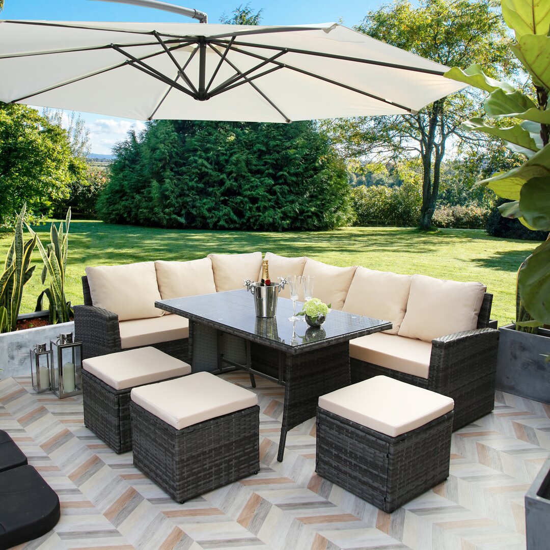 Stylish 8-seater lounge set in dark wicker with cream cushions, featuring a glass-top table and four matching stools, set under a large umbrella in a lush garden.