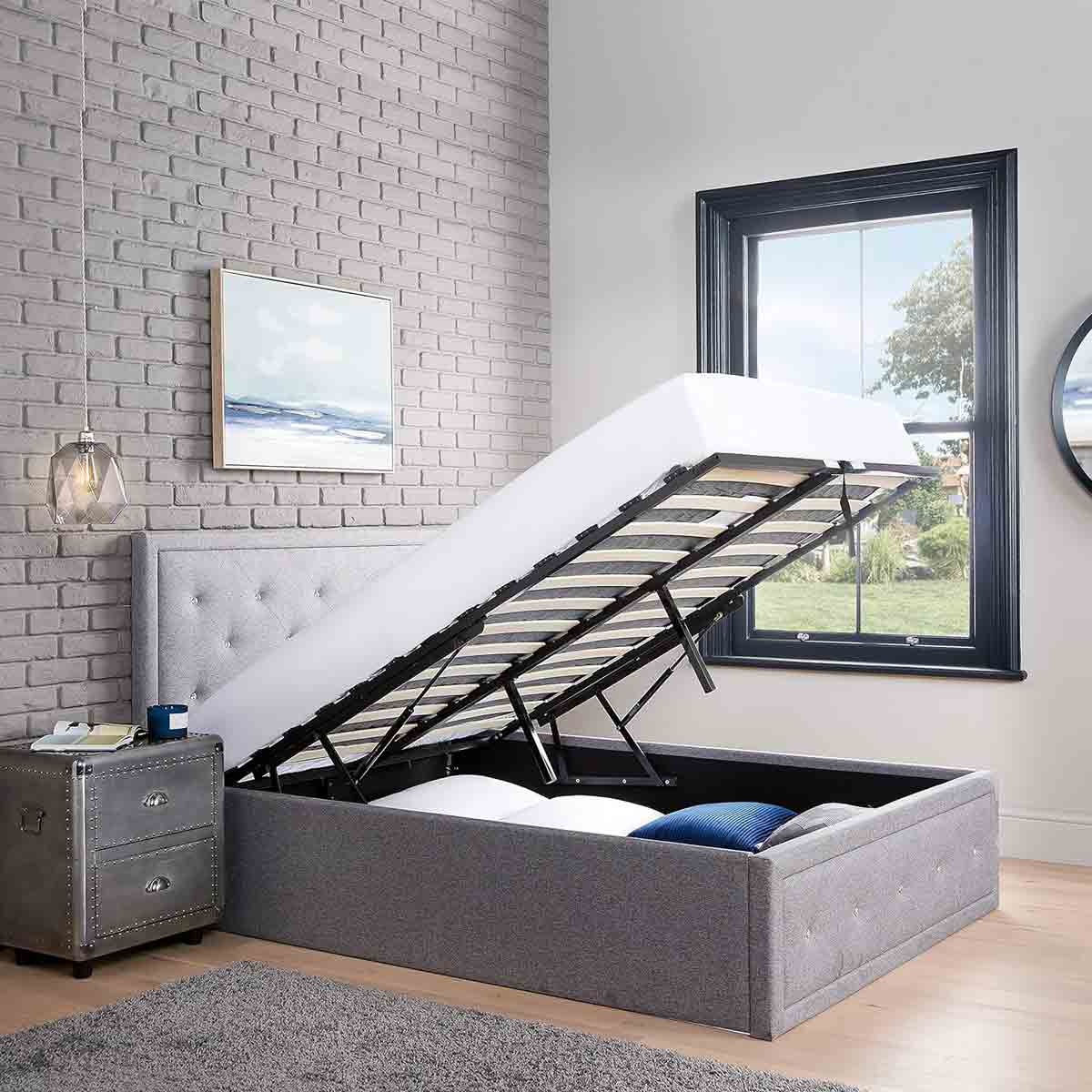 Double Ottoman storage bed in crystal grey fabric with a lift-up mattress revealing ample storage space underneath, set against a light brick wall and window backdrop.