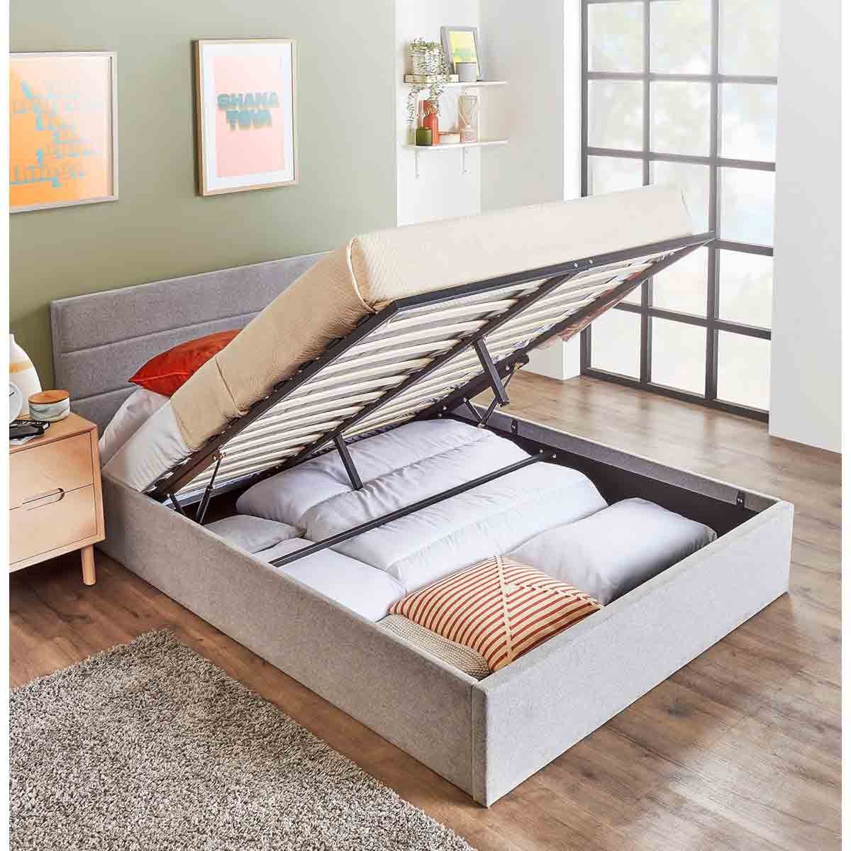 King size upholstered ottoman bed frame in grey with a lift-up storage feature, revealing ample storage space underneath, set against a light green wall and wooden floor.