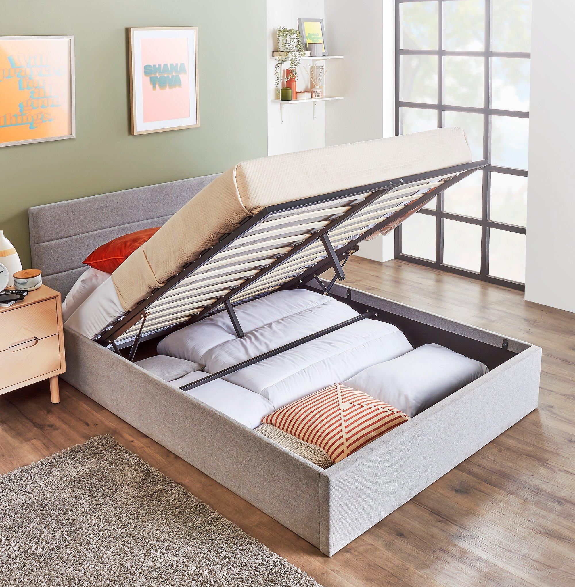 Grey upholstered double ottoman bed frame with a lift-up mattress revealing spacious storage underneath, set against a light green wall and wooden flooring.