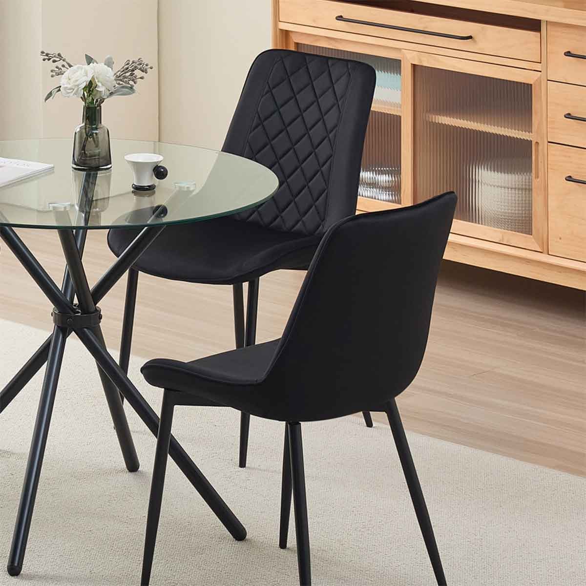 Set of 2 black velvet dining chairs with a quilted backrest, featuring sleek black legs, positioned around a glass-top table in a modern interior setting.