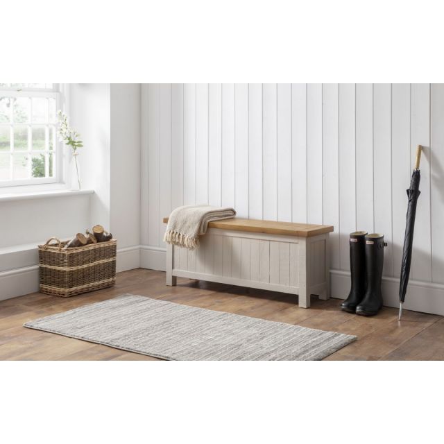 Grey wash storage bench with a wooden top, featuring slatted sides, placed against a white paneled wall, accompanied by a woven basket, umbrella, and a textured rug.