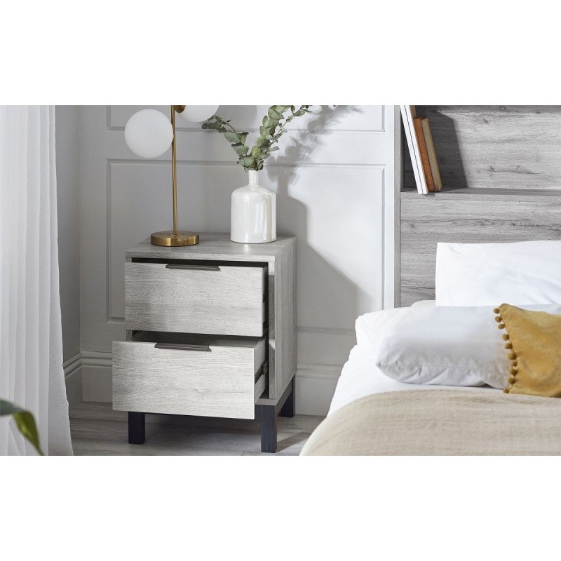 Grey oak bedside table with two drawers, sleek black handles, and tapered legs, set beside a bed with white linens and a decorative vase.