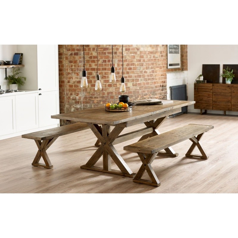 Wooden extending table with a rustic finish, featuring a sturdy X-shaped base, accompanied by two matching benches, set in a bright, modern interior with exposed brick walls.
