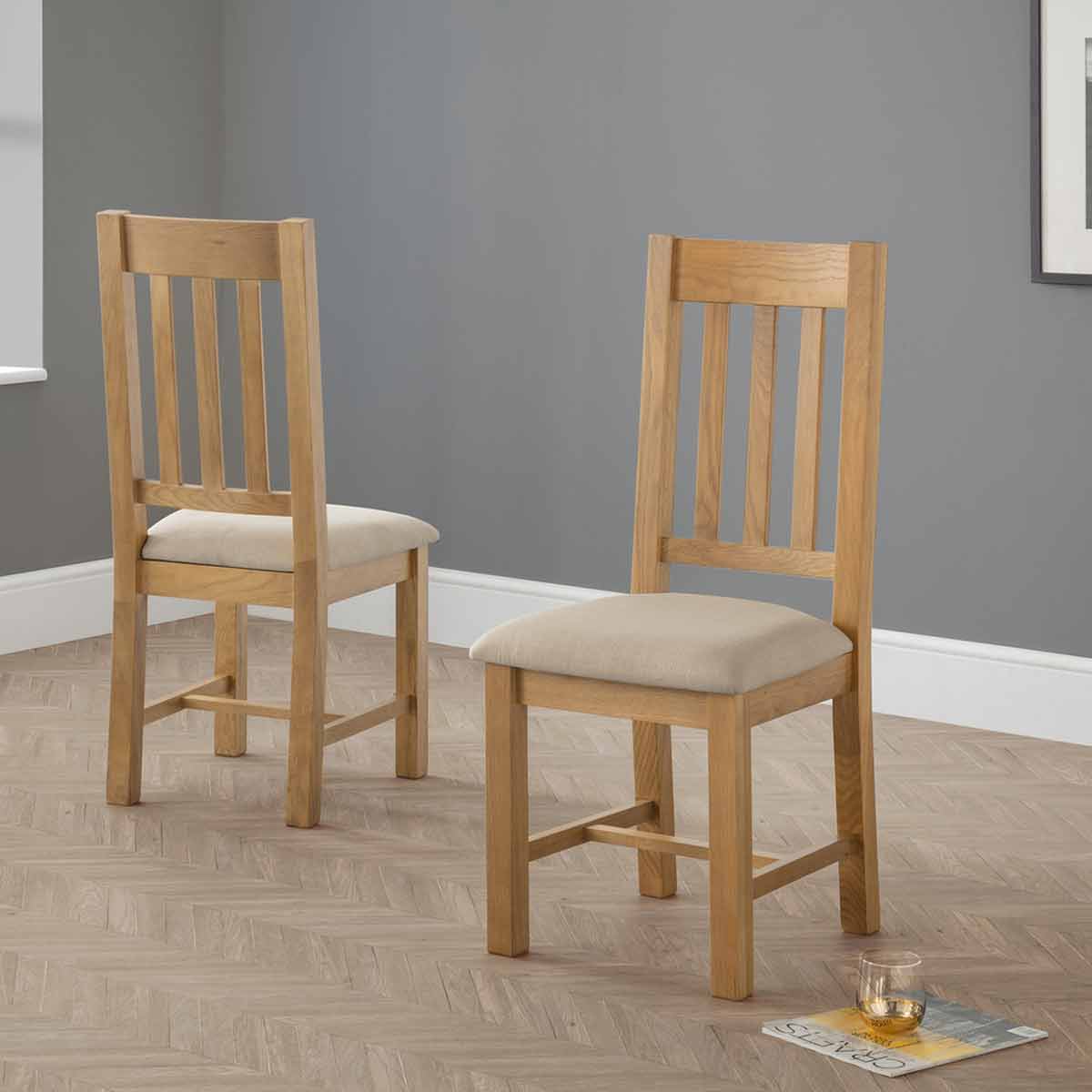 Set of 2 Hereford dining chairs featuring a natural oak finish, slatted backrest, and beige upholstered seats, positioned against a grey wall with a light wood floor.